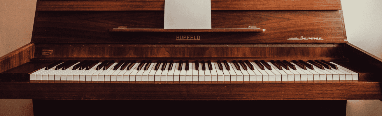 A wooden upright piano with white and black keys, featuring the brand name "Hupfeld" and model "Carmen," with a sheet of paper placed on the music stand.