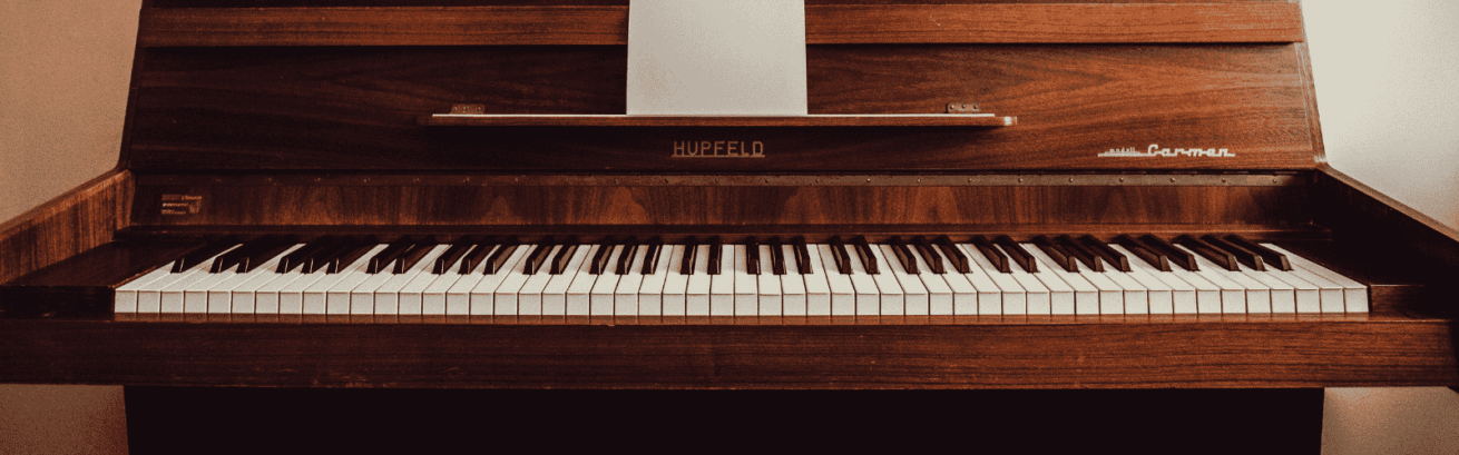 A wooden upright piano with white and black keys, featuring the brand name "Hupfeld" and model "Carmen," with a sheet of paper placed on the music stand.