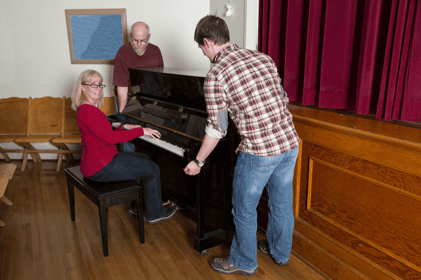 improper-disposal-of-mattress-mngmovers (1) A woman sits at an upright piano while two men prepare to move it inside a room with wooden floors, red stage curtains, and chairs along the wall.