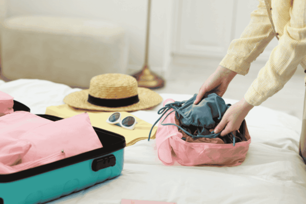 packing-cubes-mngmovers Person packing clothes into a pink packing cube beside an open suitcase on a bed, with sunglasses and a straw hat laid out for vacation travel.