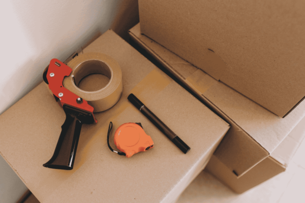 packing-supplies-marker-mngmovers Packing essentials including a tape dispenser, roll of packing tape, marker, and measuring tape placed on top of sealed cardboard moving boxes.