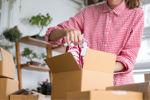 shoes-packing-supplies-mngmovers A woman in a red checkered shirt carefully packing red sneakers into a cardboard box, surrounded by other moving boxes and indoor plants in the background.