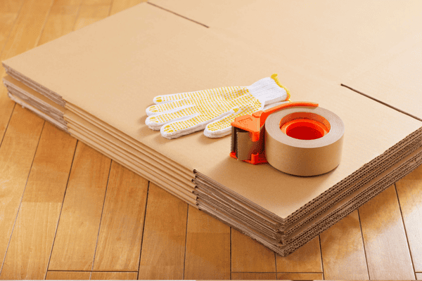 how-do-we-pack-boxes-2-mngmovers A stack of flat cardboard boxes on a wooden floor with essential packing supplies on top, including work gloves, a roll of packing tape, and a tape dispenser—ready for moving preparation.