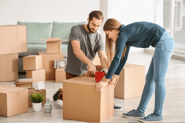 how-do-we-pack-boxes-1-mngmovers A couple sealing a moving box in a living room filled with packed boxes and household items, preparing early for an organized move.