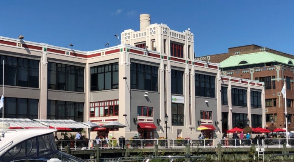 Facade of the Torpedo Factory Art Center in Alexandria, Virginia, featuring large industrial-style windows, colorful trim accents, and outdoor seating along the waterfront on a sunny day.