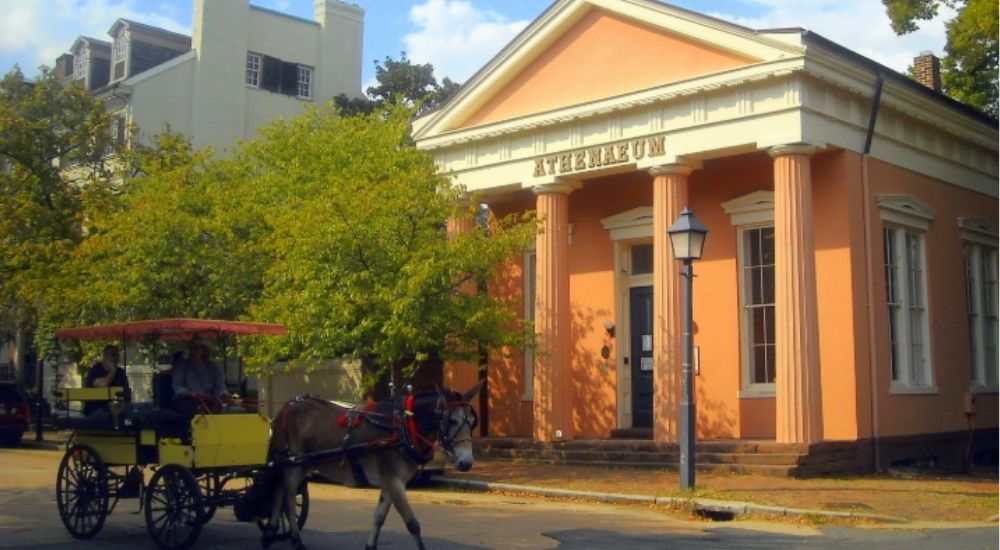 The Athenaeum museum in Alexandria, Virginia, featuring a classical Greek Revival facade with tall columns, and a horse-drawn carriage passing by on a sunny day.