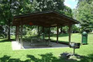 Covered picnic shelter with several tables on a concrete pad, surrounded by green grass and trees in a public park on a sunny day.