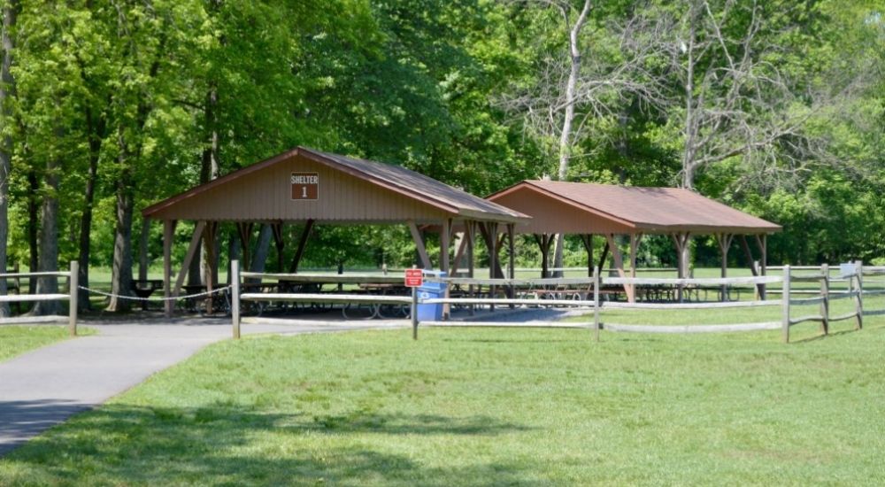 Picnic shelters at Bull Run Regional Park in Centreville, Virginia, surrounded by lush green trees and open grassy areas, with a paved path and wooden fences in the foreground.