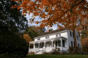 White two-story historic house with a front porch and green shutters, surrounded by trees with colorful autumn leaves.