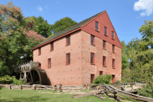 Historic red-brick gristmill with a large wooden waterwheel, red window shutters, and a split-rail fence, surrounded by trees on a sunny day.