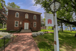 Historic red brick church building with white trim, arched windows, and a sign celebrating 250 years, surrounded by trees, flowers, and a brick walkway.