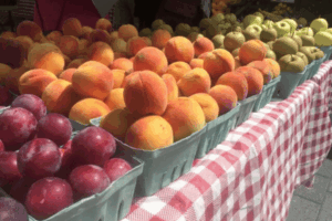 Fresh peaches, plums, and apples displayed in green cartons on a red and white checkered tablecloth at an outdoor farmers market.