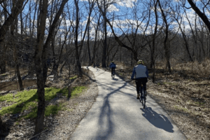 Cyclists riding along a paved trail through a wooded area with leafless trees and dappled sunlight on a clear day.
