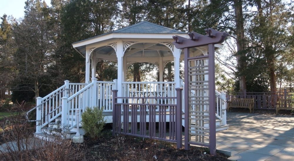 Charming white gazebo surrounded by trees and a landscaped garden at Green Spring Gardens in Virginia, with a wooden arbor at the entrance and benches nearby.