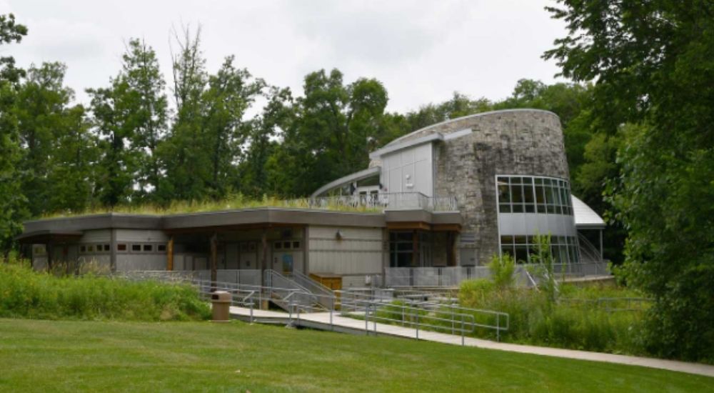 Exterior view of the Hidden Oaks Nature Center in Virginia, surrounded by lush greenery and trees, featuring a modern building with stone and glass elements and a grassy rooftop.