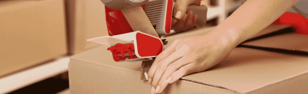 Close-up of hands using a tape dispenser to seal a cardboard box, demonstrating how we pack boxes securely and efficiently for moving.