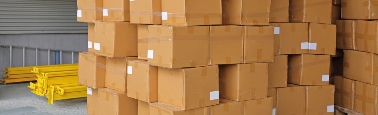 Stacks of sealed brown cardboard boxes arranged in a warehouse, ready for storage or shipping. Yellow metal shelving components are visible on the side.
