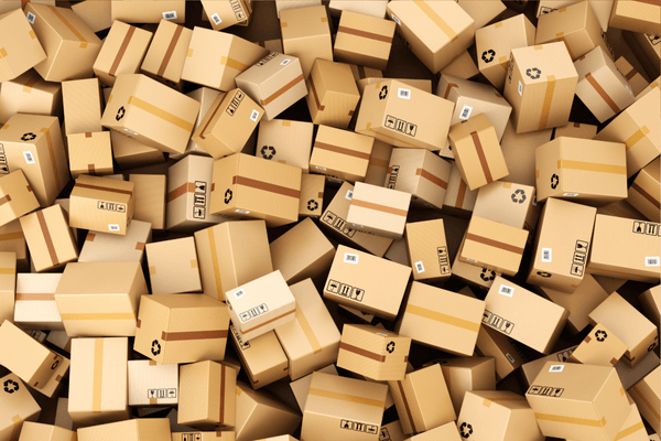 A large pile of various cardboard shipping boxes with printed labels, barcodes, and recycling symbols, scattered in a chaotic heap, symbolizing high-volume packaging or delivery.