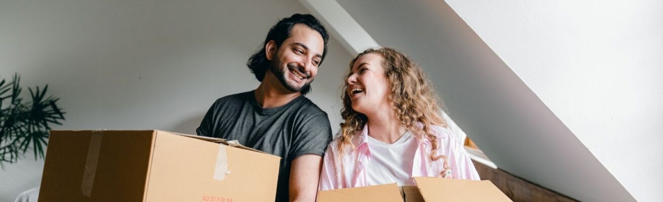 Smiling couple holding cardboard boxes in their new home, capturing a joyful moment that reflects how to make moving easier and less stressful.
