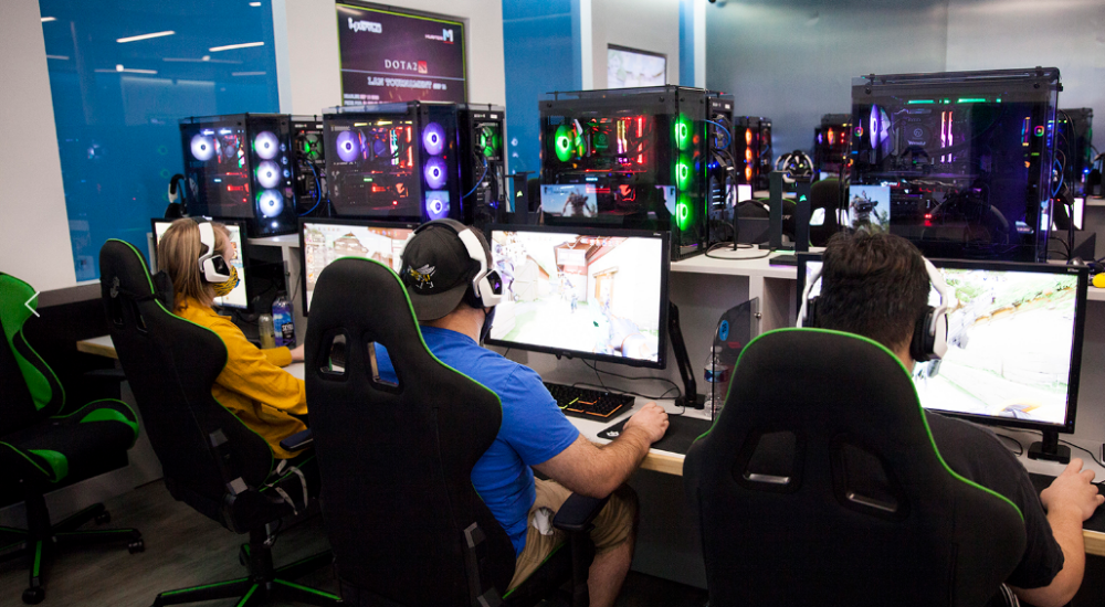 Gamers wearing headsets playing on high-performance desktop computers in a modern esports gaming center, with RGB-lit PC towers and ergonomic gaming chairs.
