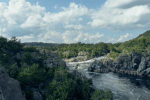 Scenic view of Great Falls on the Potomac River, with rocky cliffs, lush green trees, and a cloudy blue sky on a summer day.