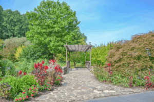 A stone pathway leads to a wooden arbor surrounded by colorful flowers and lush greenery at Meadowlark Botanical Gardens, with vibrant landscaping and tall trees under a clear blue sky.