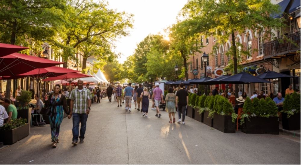 Busy pedestrian scene on King Street in Old Town Alexandria, VA, with people walking, outdoor dining under umbrellas, and historic brick buildings lined with shops and trees at sunset.