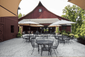 Outdoor seating at Paradise Springs Winery in Clifton VA, featuring a red barn-style tasting room, patio tables with umbrellas, and scenic vineyard surroundings.