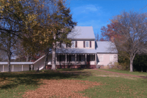 Historic two-story colonial house with a front porch and white shutters, surrounded by trees with autumn foliage and a grassy lawn covered in fallen leaves.