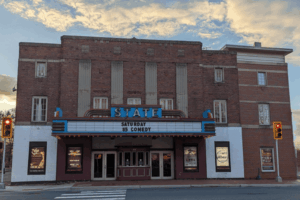 Historic brick movie theater with a vintage marquee reading "Saturday $5 Comedy," flanked by poster displays and lit by a warm evening sky.