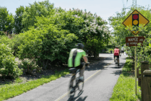 Cyclists ride along a sunny, tree-lined section of the Washington & Old Dominion Trail in Vienna, Virginia, near a sign for Maple Avenue and a traffic signal warning.