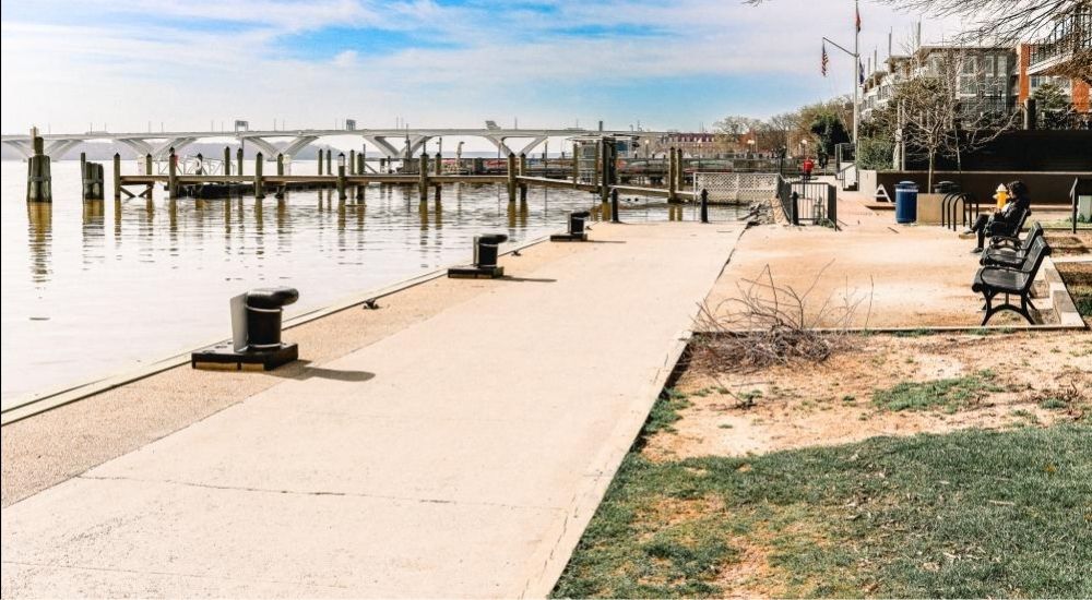 Scenic view of Waterfront Park in Alexandria, Virginia, featuring a quiet promenade along the Potomac River with benches, a dock, and the Woodrow Wilson Bridge in the background on a clear day.