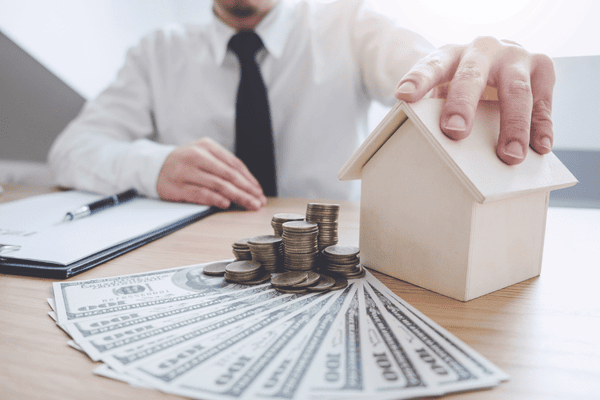 why-do-people-move-mngmovers-1 Man in a suit with a model house, coins, and dollar bills on a desk, illustrating how rising housing costs can impact the decision to move.