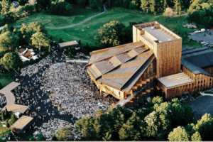 ChatGPT said: A large crowd gathers outside the distinctive hexagonal roof structure of the Filene Center at Wolf Trap National Park for the Performing Arts, surrounded by lush green trees and open parkland.